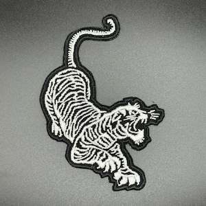 May include: White embroidered tiger patch with black outline. The tiger is leaping with its mouth open and its tail curled up.
