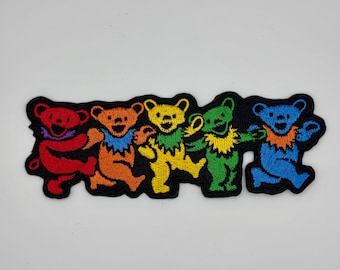 May include: Five colorful dancing bears in a row. The bears are red, orange, yellow, green, and blue. The bears are embroidered on a black fabric patch.