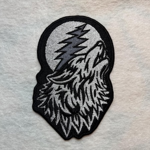 May include: Embroidered patch featuring a howling wolf against a full moon and a stylized tree. The design uses black, gray, and white thread. The patch has a black border and is likely for clothing or accessories.