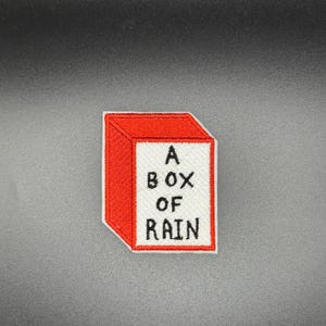 May include: Embroidered patch featuring a red box with the words "A BOX OF RAIN" in black lettering on a white background. The patch has a red border and is likely designed to be ironed or sewn onto fabric.