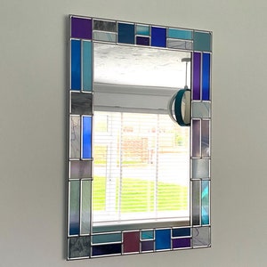 May include: A rectangular mirror with a stained glass frame. The frame is made of small pieces of glass in shades of blue, purple, and green. The mirror is reflecting a window with blinds and a green lawn.