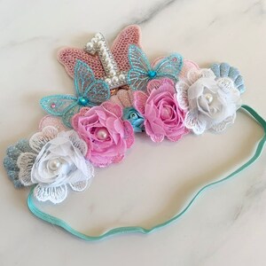 May include: A light blue headband with a white, pink, and blue flower crown. The crown features a sparkly silver number one with a pearl center and a butterfly design.