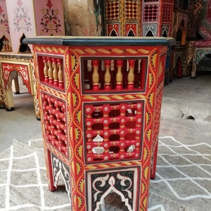 May include: A hand-painted, red wooden side table with intricate geometric and floral designs. The table features a latticed front, decorative balusters, and black and white accents. Other similar tables are visible in the background.