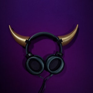 May include: Black headphones with gold horns attached to the top. The headphones are on a purple background.