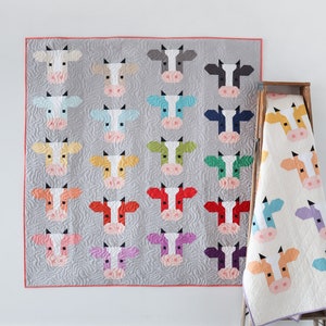 Puede incluir: Una colcha con un diseño de patchwork de caras de vacas de colores. La colcha está hecha de cuadrados con caras de vacas de diferentes colores en cada cuadrado. La colcha está colgada en una pared blanca.