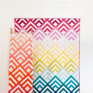 May include: Two colourful quilts with a chevron pattern in shades of pink, orange, yellow, green, blue, and white. The quilts are hanging on a wooden ladder against a white wall.