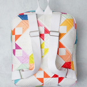 May include: A colourful patchwork backpack with white adjustable straps and silver metal buckles. The bag features a white background with geometric shapes in red, orange, yellow, blue, and pink. The backpack is designed for carrying items, and is quilted.