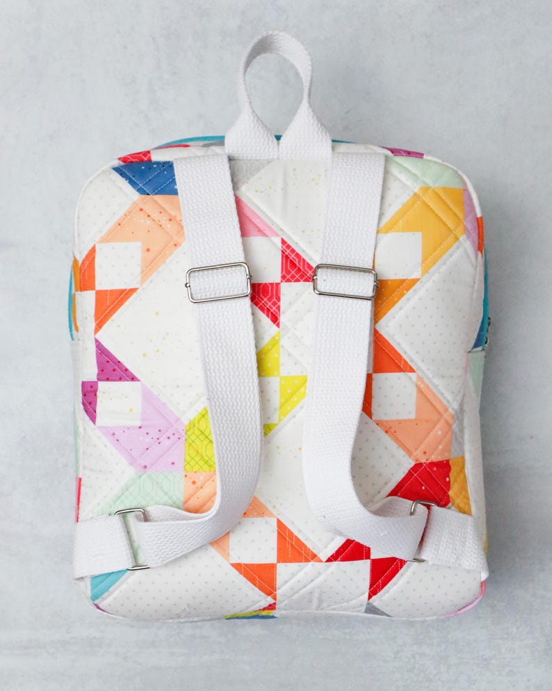 May include: A colourful patchwork backpack with white adjustable straps and silver metal buckles. The bag features a white background with geometric shapes in red, orange, yellow, blue, and pink. The backpack is designed for carrying items, and is quilted.