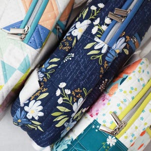May include: Close-up of several quilted fabric bags with silver zips. One bag features a navy blue floral pattern, while others display colourful geometric and floral designs. The bags are rectangular and appear to be padded.