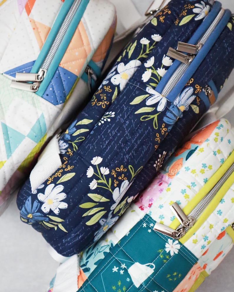May include: Close-up of several quilted fabric bags with silver zips. One bag features a navy blue floral pattern, while others display colourful geometric and floral designs. The bags are rectangular and appear to be padded.
