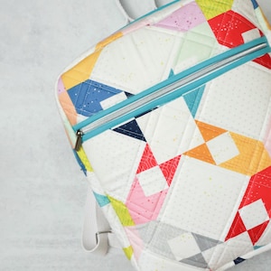 May include: A colourful patchwork backpack. The bag has a white base with geometric shapes in red, pink, yellow, blue, and orange. It features a teal zip and white straps. The bag is quilted.