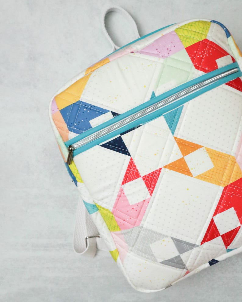 May include: A colourful patchwork backpack. The bag has a white base with geometric shapes in red, pink, yellow, blue, and orange. It features a teal zip and white straps. The bag is quilted.