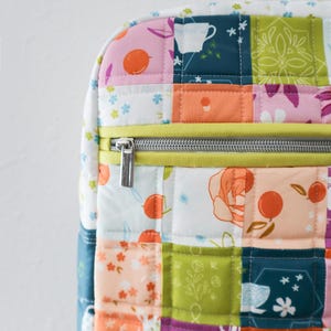 May include: A patchwork backpack with a variety of floral and fruit patterns in pink, orange, green, and blue. The bag has a yellow zip and a silver zip pull. The bag has a white handle. The bag is quilted.