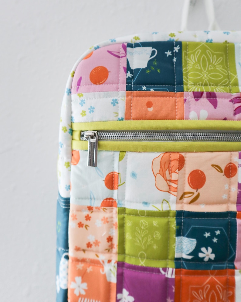 May include: A patchwork backpack with a variety of floral and fruit patterns in pink, orange, green, and blue. The bag has a yellow zip and a silver zip pull. The bag has a white handle. The bag is quilted.