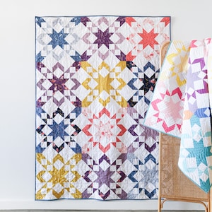 May include: A colorful quilt with a starburst pattern in shades of blue, purple, pink, and yellow. The quilt is made up of individual squares with different patterns and colors. The quilt is hanging on a white wall.