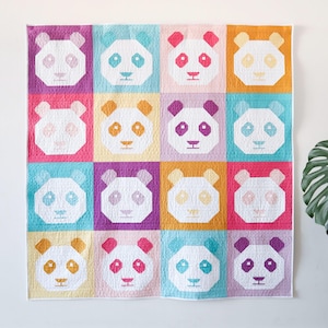 Op de afbeelding: Een kleurrijk quilt met een herhaald patroon van panda gezichten. Elk panda gezicht is geplaatst tegen een ander pastelkleurig vierkant, waaronder roze, blauw, paars en oranje. De panda's hebben witte gezichten met zwarte ogen en oren en een kleine rode neus.