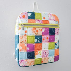 May include: A colorful patchwork backpack with a quilted design. The backpack features a variety of patterned squares in shades of pink, orange, green, and blue. It has a yellow zipper and a white handle. The backpack is hanging on a white wall.