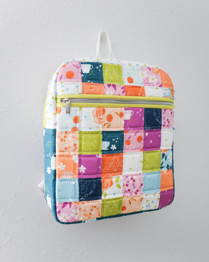 May include: A patchwork backpack in a quilted design. The bag features squares in pink, orange, green, and blue. It has a yellow zip and a white handle. The backpack is hanging on a white wall. The dimensions are approximately 30 cm in height and 25 cm in width.