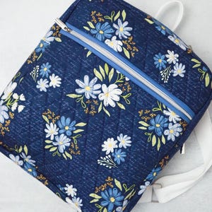 May include: A navy blue quilted backpack with a floral pattern of white and blue flowers. It features a silver zip, light blue trim, and white straps. The design includes a subtle script-like background.
