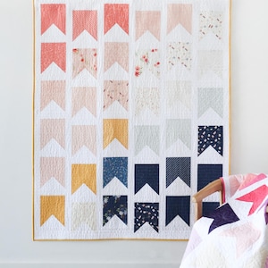 May include: A white quilt with a pattern of colorful pennants. The pennants are in shades of pink, yellow, blue, and gray. The quilt is hanging on a white wall.