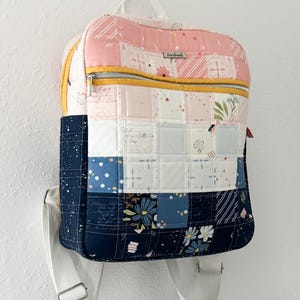 May include: A handmade, quilted backpack with a patchwork design. The top section is pink with floral and striped patterns, while the middle section features white and blue squares. The bottom section is navy blue with floral patterns. A yellow zip and white straps complete the design.