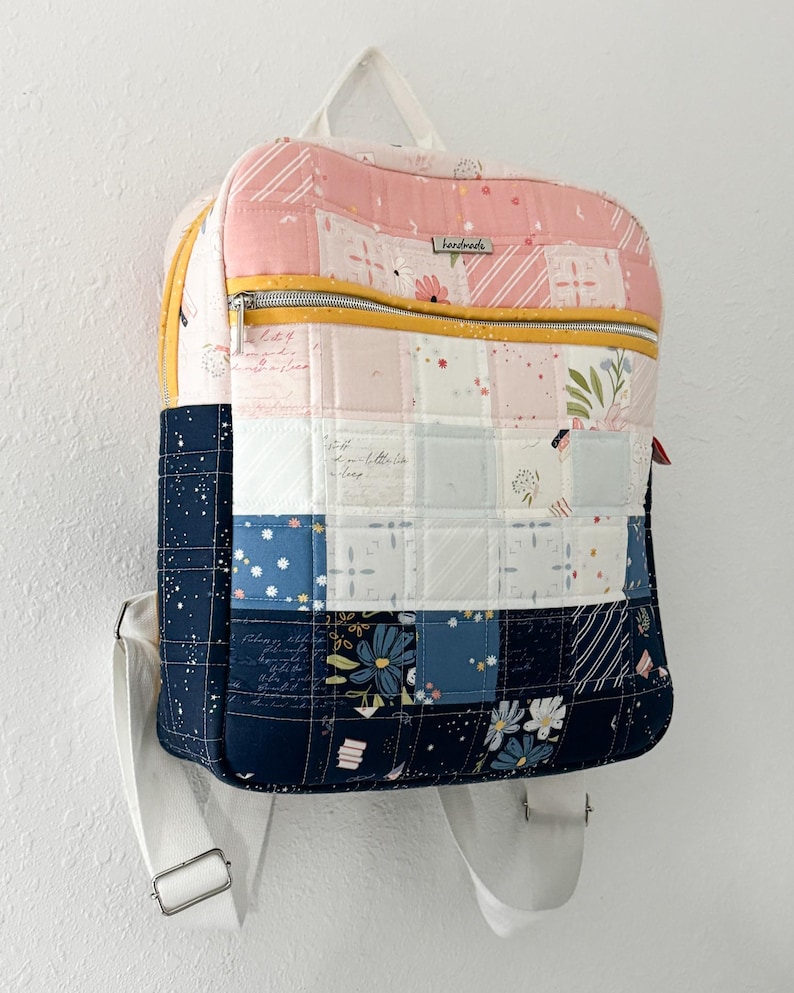 May include: A handmade, quilted backpack with a patchwork design. The top section is pink with floral and striped patterns, while the middle section features white and blue squares. The bottom section is navy blue with floral patterns. A yellow zip and white straps complete the design.