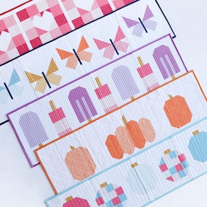 May include: Five different fabric table runners with various designs. The designs include pink and white gingham, butterflies, popsicles, pumpkins, and ornaments. The runners are all white with a white background and are trimmed in different colors.
