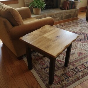 May include: A square side table featuring a natural live edge light wood top with visible grain patterns and four straight, dark brown wooden legs. The table surface has a smooth finish and a distinct organic edge profile on all four sides.