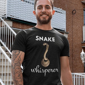 Könnte beinhalten: Ein schwarzes T-Shirt mit weißem Text, der "SNAKE whisperer" lautet. Über dem Text ist eine braune Schlange mit erhobenem Kopf abgebildet.