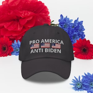 Puede incluir: Una gorra de béisbol negra con el texto "PRO AMERICA ANTI BIDEN" bordado en la parte delantera. Tres pequeñas banderas americanas están bordadas encima del texto.