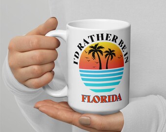 Puede incluir: Taza de cerámica blanca con un gráfico de puesta de sol y palmeras. El texto "I'd rather be in Florida" está impreso en la taza.