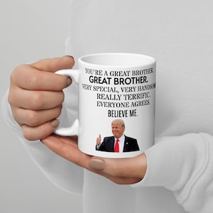 May include: White ceramic mug with a black and white image of a man with a thumbs up gesture. The text on the mug reads "You're a great brother. Great brother. Very special, very handsome. Really terrific. Everyone agrees. Believe me."