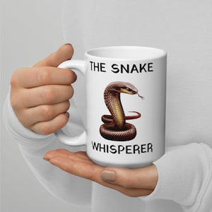 Könnte beinhalten: Eine weiße Keramiktasse mit einem schwarzen und braunen Schlangenmotiv und dem Text "The Snake Whisperer".