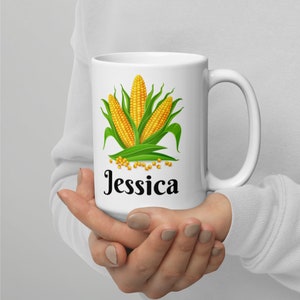 Op de afbeelding: Witte keramische mok met een ontwerp van drie maïsoren en de naam "Jessica" in het zwart gedrukt.