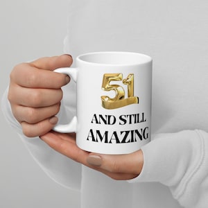 May include: White ceramic coffee mug with a black and gold design. The mug features the number 51 in gold with a black outline and the text "And Still Amazing".