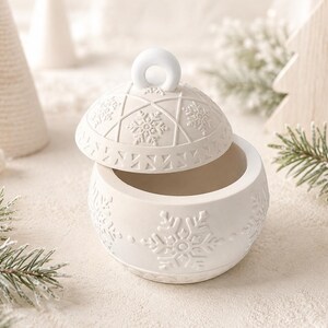 Könnte beinhalten: Ein mattweißer Keramikbehälter mit Deckel in Form einer runden Weihnachtskugel. Die Außenseite ist mit geprägten Schneeflockenmustern und geometrischen Linien verziert. Der Deckel verfügt über einen kleinen runden Griff an der Oberseite.