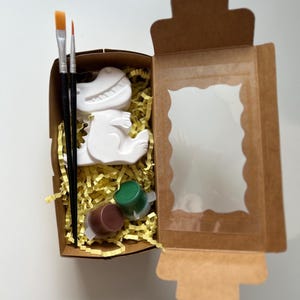 Könnte beinhalten: Ein Bastelset in einer braunen Pappschachtel mit einer weißen Dinosaurierfigur, Pinseln und Farbtöpfen. Die Schachtel ist mit gelben Papierschnipseln gefüllt. Das Set enthält braune und grüne Farbe.