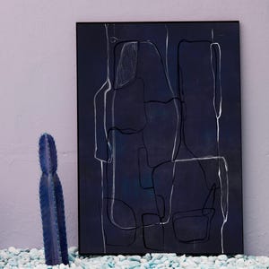 May include: A framed abstract art print with a dark blue background and white line art. Two blue cacti stand in front of a light purple wall and white pebbles. The artwork has a modern, minimalist aesthetic.