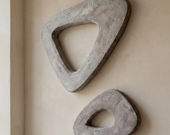 May include: Two gray, abstract wall sculptures with a triangular shape and a hole in the center. The sculptures are mounted on a light beige wall, creating a modern, minimalist aesthetic. The larger sculpture is above the smaller one.
