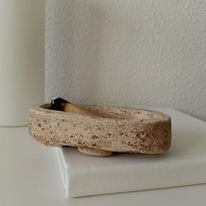 May include: A beige, oval incense holder with a textured surface. A lit incense stick rests inside the holder. The holder sits on a white surface, with a white candle in the background. The incense holder is a decorative item.