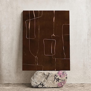 Può includere: Opera d'arte astratta. Una tela rettangolare marrone scuro con disegni lineari beige chiaro poggia su una pietra grigia strutturata con graffiti e accenti floreali rosa. Lo sfondo è un muro grigio chiaro neutro.