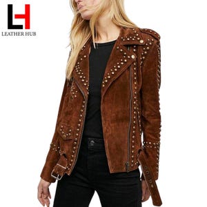 May include: A brown suede jacket adorned with gold-tone studs. It has an asymmetrical zip, a notched collar, and a waist belt. The cuffs have zip detailing. The Leather Hub logo is visible in the top left corner. The jacket is a stylish outerwear piece.