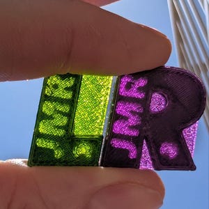 May include: Close-up of a 3D printed "LR" keychain. The "L" is green and the "R" is purple.