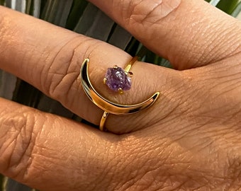 May include: Gold crescent moon ring with a purple gemstone. The ring is on a finger.