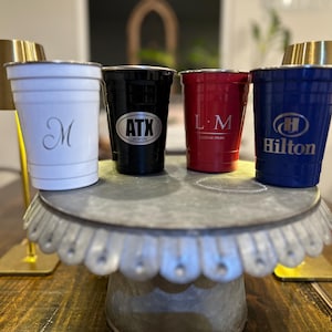 May include: Four metal cups in white, black, red, and blue, each with a different logo or initial. The cups are arranged on a decorative metal stand. The white cup has a cursive "M", the black cup has "ATX", the red cup has "L.M", and the blue cup has the Hilton logo.