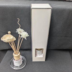 May include: An aromatherapy diffuser set with a white rectangular box and a small glass bottle with a label. The set includes a small glass bottle with a wooden cap, a clear glass saucer, and decorative white flowers.