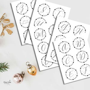 May include: Several white cards displaying cursive alphabet letters, each within a wreath of leaves and hearts. The words "Merry Christmas" are also visible. Christmas baubles are in the foreground, adding a festive touch.