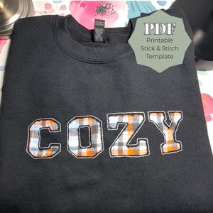 May include: Black sweatshirt with the word "COZY" appliqued across the front. The letters are filled with a plaid pattern in orange, white, and blue. A tag in the upper right corner reads "PDF Printable Stick & Stitch Template".