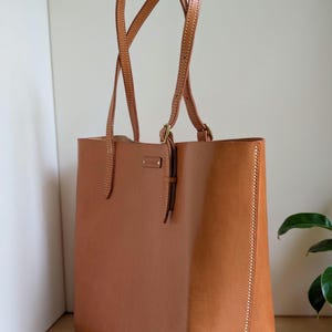 May include: A tan leather tote bag featuring two long shoulder straps with adjustable brass buckles, a rectangular body, and visible light-colored stitching along the side seams. A small rectangular leather tag is attached near the top center.