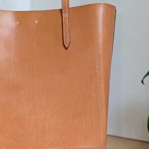 May include: A tan leather tote bag featuring visible white stitching along the edges and a vertical strap attachment. The surface displays natural grain textures and two small metal rivets near the top edge of the bag's main body.